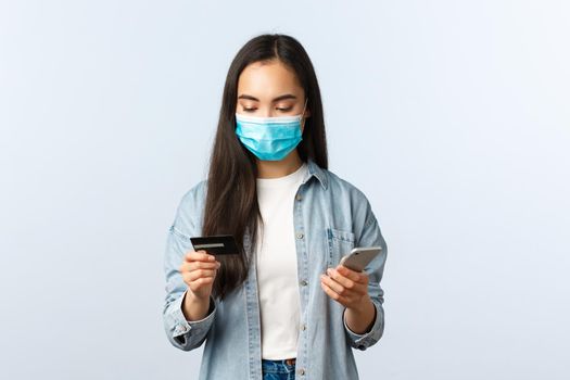 Social distancing lifestyle, covid-19 pandemic and contactless shopping concept. Attractive stylish asian girl in medical mask looking at credit card while making order on mobile application.