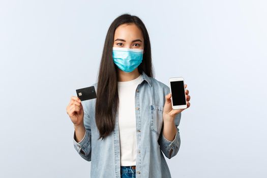 Social distancing lifestyle, covid-19 pandemic and contactless shopping concept. Young pretty asian woman in medical mask showing mobile phone screen and credit card, buying online.