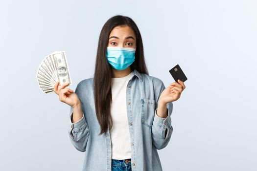 Social distancing lifestyle, covid-19 pandemic business and employement concept. Indecisive cute asian girl in medical mask shrugging shoulders as holding cash dollars and credit card.