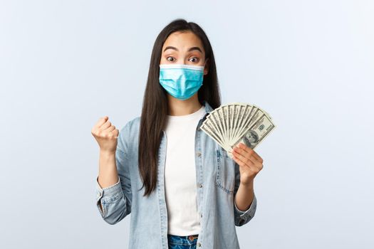 Social distancing lifestyle, covid-19 pandemic business and employement concept. Excited happy asian woman winning lottery, wear medical mask and rejoicing, show money, holding cash.