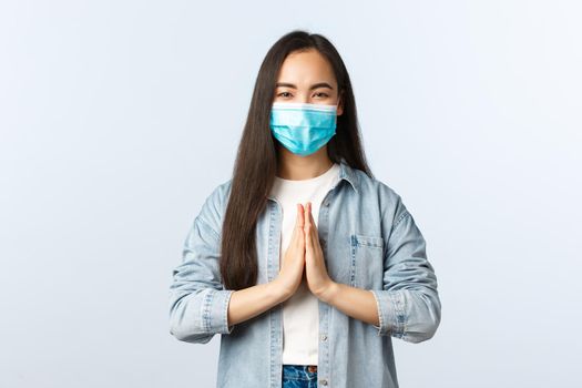 Social distancing lifestyle, covid-19 pandemic everyday life and leisure concept. Cheerful carefree asian woman in medical mask say namaste, hold hands in plead gesture and smiling.