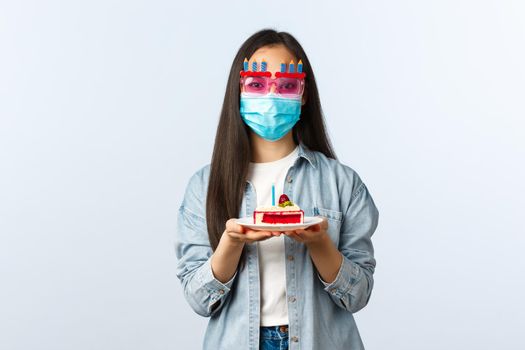 Social distancing lifestyle, covid-19 pandemic, celebrating holidays during coronavirus concept. Cute female friend in medical mask and party glasses giving b-day girl birthday cake.
