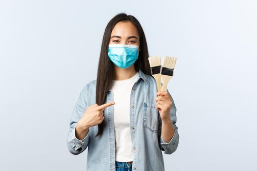 Social distancing lifestyle, covid-19 pandemic, self-isolation hobbies and leisure concept. Cute artistic and creative asian female student plan rebuilding house, showing paint brushes, wear mask.