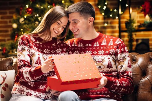 Happy man is making christmas gift to his beloved woman. Portrait of a romantic couple opening a present gift box in the evening near decorated xmas tree. Concept of holidays, romance, surprise.
