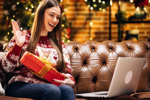 Cheerful young caucasian lady sit on couch chat in video conference app on laptop show gift box to webcam. Excited millennial lady call good friend by pc greet with New Year. Gifts from Santa.