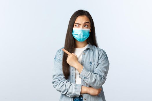 Social distancing lifestyle, covid-19 pandemic everyday life concept. Skeptical unsure young asian girl in medical mask, looking and pointing finger upper left corner with doubtful uncertain face.