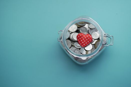 Red heart and coins in a jar on light green background .