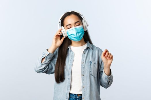 Social distancing lifestyle, covid-19 pandemic and self-isolation leisure concept. Carefree dreamy asian girl in medical mask enjoying listening music in wireless headphones, close eyes relaxed.