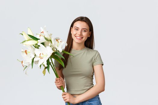 Happy good-looking young caucasian woman receive bouquet delivery, smiling at camera as holding flowers. Girl buy lilies for her home, standing white background pleased.