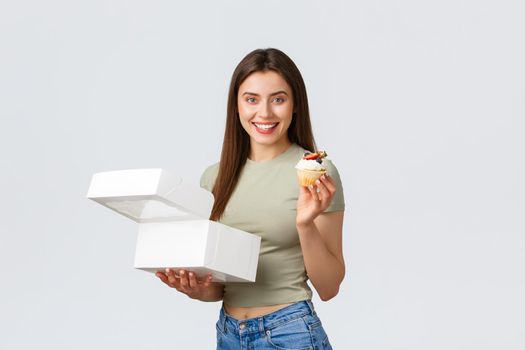 Delivery, lifestyle and food concept. Cheerful happy pretty woman standing with opened box from pastry shop and cupcake, smiling at camera after taking order from courier, eating tasty food.