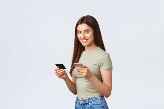 Online shopping, home lifestyle and people concept. Smiling attractive woman in casual clothes booking flight with smartphone, holding credit card and mobile phone to pay for delivery, place order.