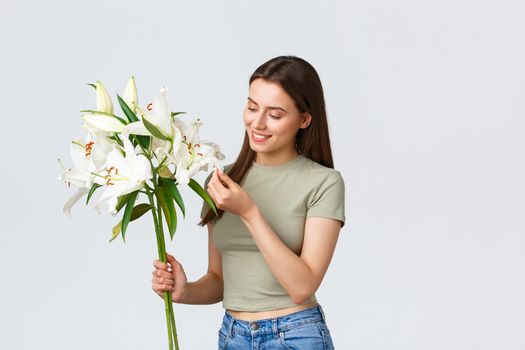 Smiling good-looking caucasian woman receive flowers delivery from florist shop, looking happy at bouquet white lilies. Girl dreamy touching leaf and thinking of boyfriend who made romantic gesture.