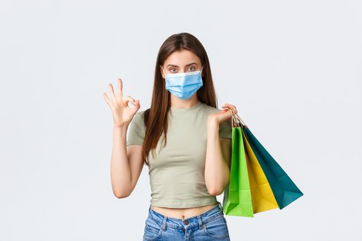 Social distancing, coronavirus and shopping, fashion concept. Stylish good-looking woman in medical mask holding shopping bags and show okay sign as recommend store in mall, white background.