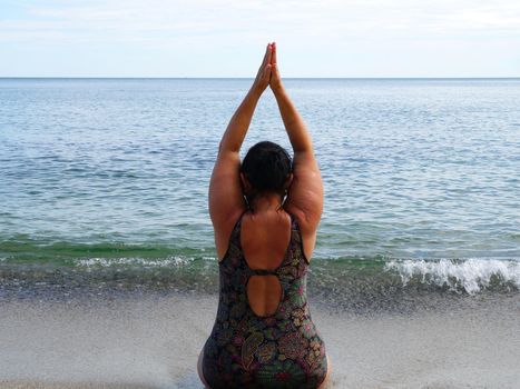 woman in a swimsuit performs yoga while sitting on the shore at the water's edge, back view