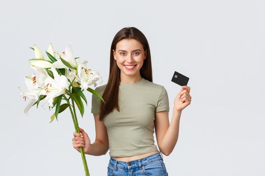 Happy tender young woman ordered bouquet of flowers delivery with credit card, smiling pleased and holding beautiful white lilies. Girl paying for purchase in internet, standing white background.