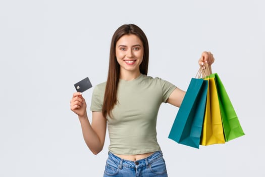 Shopping mall, lifestyle and fashion concept. Excited girl buying new clothes for summer vacation, smiling and showing credit card with bags fom stores, white background.