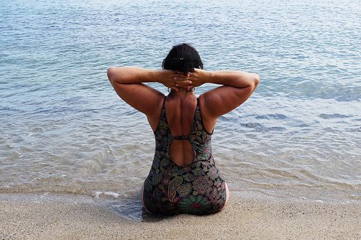 woman in a swimsuit performs yoga while sitting on the shore at the water's edge, back view