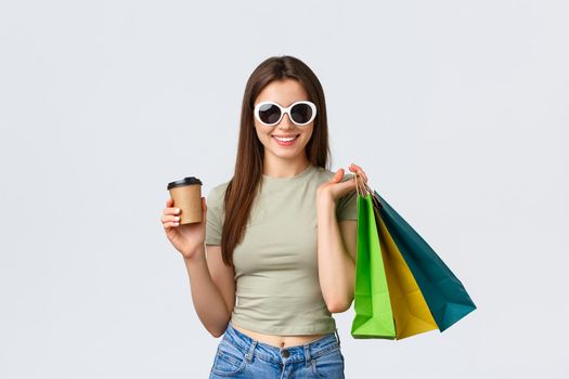 Shopping mall, lifestyle, tourism and fashion concept. Stylish carefree woman in sunglasses, drinking coffee and purchase clothes, holding takeaway cup and shop bags.