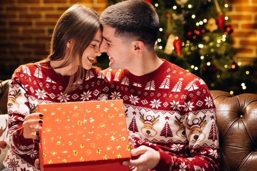 Portrait of a romantic couple opening a present gift box in the evening near decorated xmas tree. Concept of holidays, romance, surprise. Happy man is making christmas gift to his beloved woman.