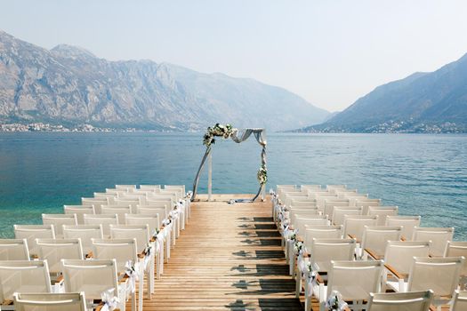 Wedding arch stands in front of a row of chairs on a wooden pier overlooking the mountains. High quality photo