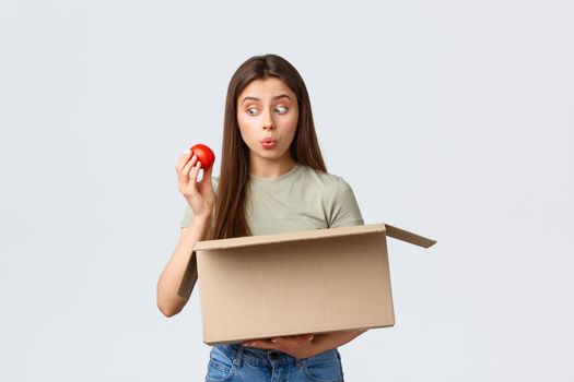 Online home delivery, internet orders and grocery shopping concept. Excited cute female customer receive groceries from internet store, take-out tomato and smiling amused.