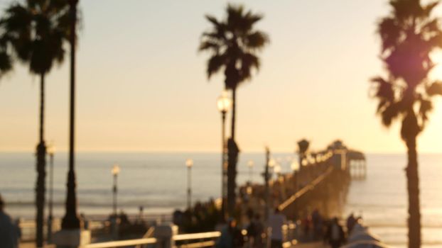 People walking, wooden pier in California USA. Oceanside waterfront vacations tourist resort. Ocean beach summertime sunset atmosphere. Blurred crowd strolling seaside boardwalk. Defocused palm trees.
