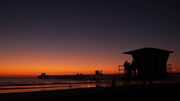 Young teen girls silhouettes near lifeguard tower, friends on pacific ocean beach, sunset dusk in Oceanside, California USA. Unrecognizable teenagers, people and twilight gradient purple violet sky.