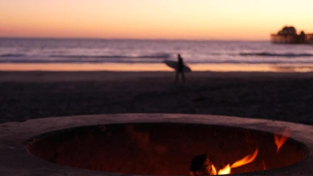 Campfire pit by Oceanside pier, California USA. Camp fire burning on ocean beach, bonfire flame in cement ring place for bbq, sea water waves. Romantic evening twilight dusk sky. Surfer with surfboard