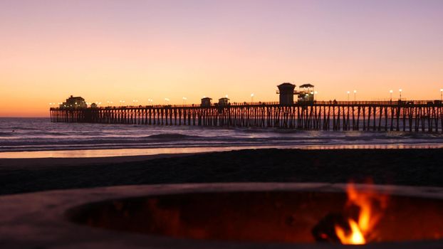 Campfire pit by Oceanside pier, California USA. Camp fire burning on ocean beach, bonfire flame in cement ring place for bbq, sea water waves. Romantic evening twilight sky, dusk after summer sunset.