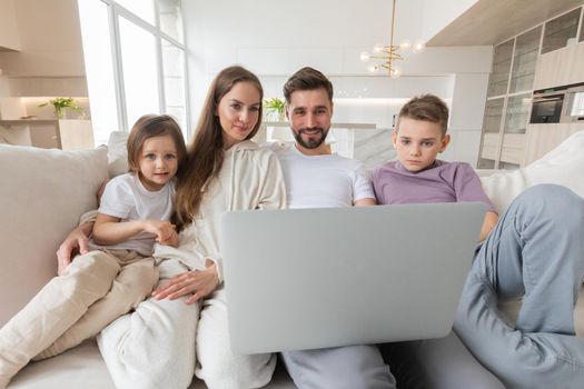 Portrait of family of parents with their two children relaxing in living room sitting together browsing internet on laptop computer
