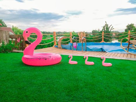 Big inflatable and three small flamingos on the green grass in front of the pool. Coasters for the pool. The concept of summer spending time. Beach summer composition