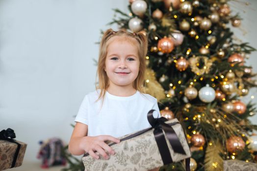 Very nice charming little girl blonde holds a gift on a background of Christmas trees in the interior of the house