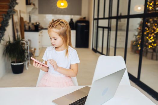 Little kid sit at desk with laptop hold mobile phone at home