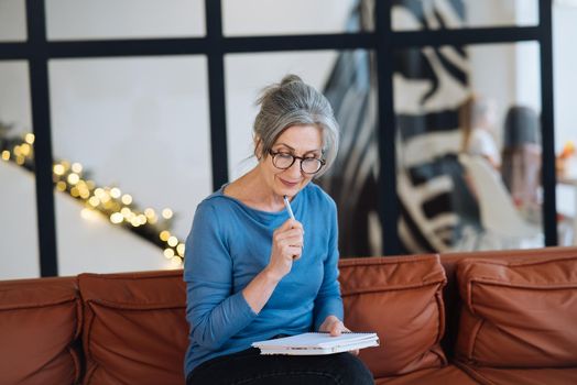 Happy senior woman in glasses writing to notebook or diary at home