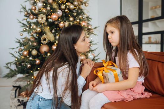 Young mother calming down her sad infant daughter sitting on the Christmas tree background