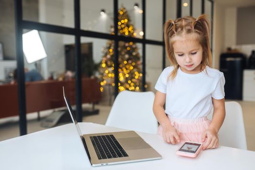 Little kid sit at desk with laptop hold mobile phone at home