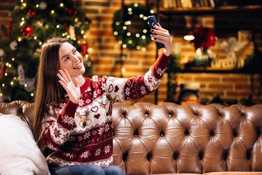 Young caucasian woman using smart phone video call talking with friend or boyfriend. Christmas tree decorated with ornament in living room at home. Christmas and New Year holiday festival