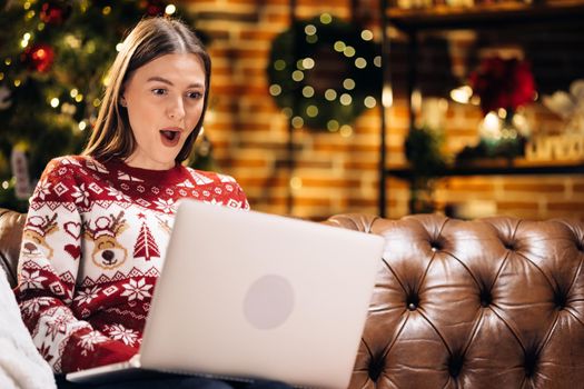 Young pretty cheerful happy girl look laptop xmas win inside home. Woman sitting on sofa with laptop celebrating victory with christmas decorations indoors. Screaming for success. Celebration concept.
