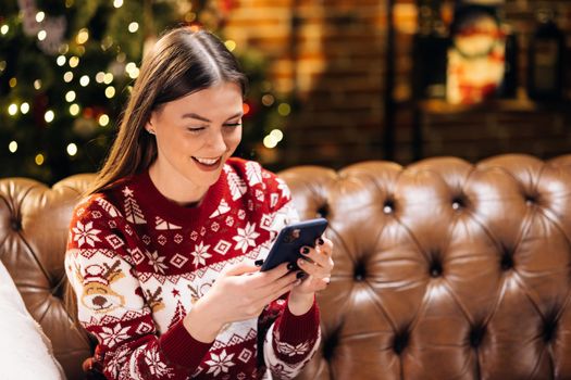 Cheerful young pretty woman using smartphone, playing online games, communicating in social network sending Christmas greetings for friends or family online resting on sofa near Christmas tree at home