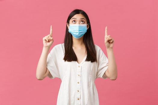 Covid-19, social distancing, virus and lifestyle concept. Curious pretty asian girl in medical mask and summer dress, looking and pointing fingers up at promo, advertisement, pink background.