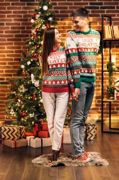 Passionate romantic couple is spending time together before New Year near beautiful Christmas tree at home. Young woman and handsome man are having dressed in winter Xmas soks in cozy home interior.