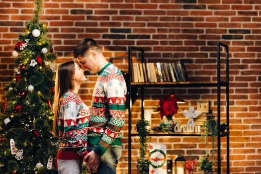 Passionate romantic caucasian couple in sweaters are spending time together before New Year at home. Tender couple hugging on the background of shiny garland.