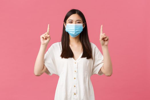 Covid-19, social distancing, virus and lifestyle concept. Cheerful smiling woman in stylish white dress, summer outfit and medical mask, pointing fingers up, promoting advertisement, pink background.