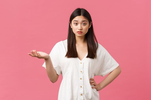 Beauty, people emotions and summer leisure concept. Confused and puzzled asian girl in white dress, raise hand up and shrugging, cant understand what you want, pink background.