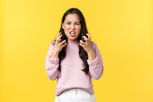Lifestyle, emotions and advertisement concept. Outraged and pissed-off asian girl looking mad and aggressive clench fists, want kill someone, grimacing angry, standing yellow background.