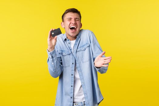 Enthusiastic handsome blond guy laughing joyfully, showing credit card amazed, rejoicing over awesome service, winning money, got paid, standing upbeat over yellow background.