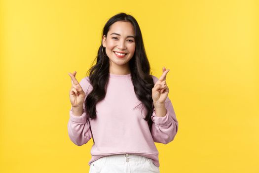People emotions, lifestyle and fashion concept. Hopeful and optimistic cute asian girl having high hopes, cross fingers good luck and smiling, making wish, standing yellow background.