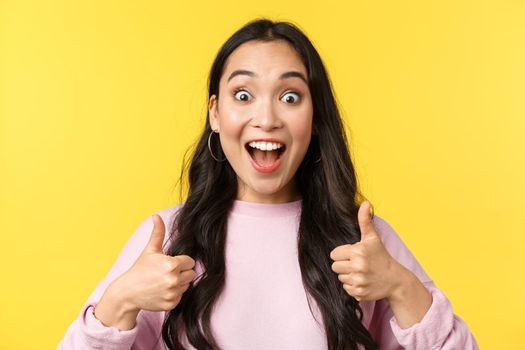 People emotions, lifestyle leisure and beauty concept. Close-up of enthusiastic and amazed, smiling asian girl hear awesome news, show thumbs-up in approval, like idea, yellow background.