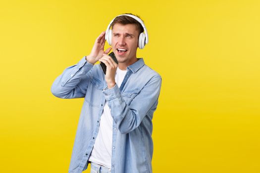 Lifestyle, summer holidays, technology concept. Happy good-looking blond guy in casual outfit, playing karaoke app, singing song into mobile phone and wearing headphones, yellow background.