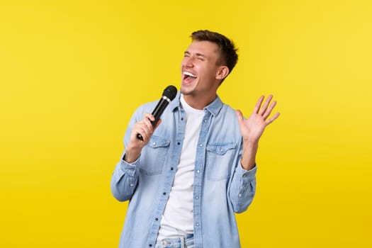Lifestyle, people emotions and summer leisure concept. Happy carefree handsome man singing with passion, holding microphone, perform favorite song in karaoke, yellow background.